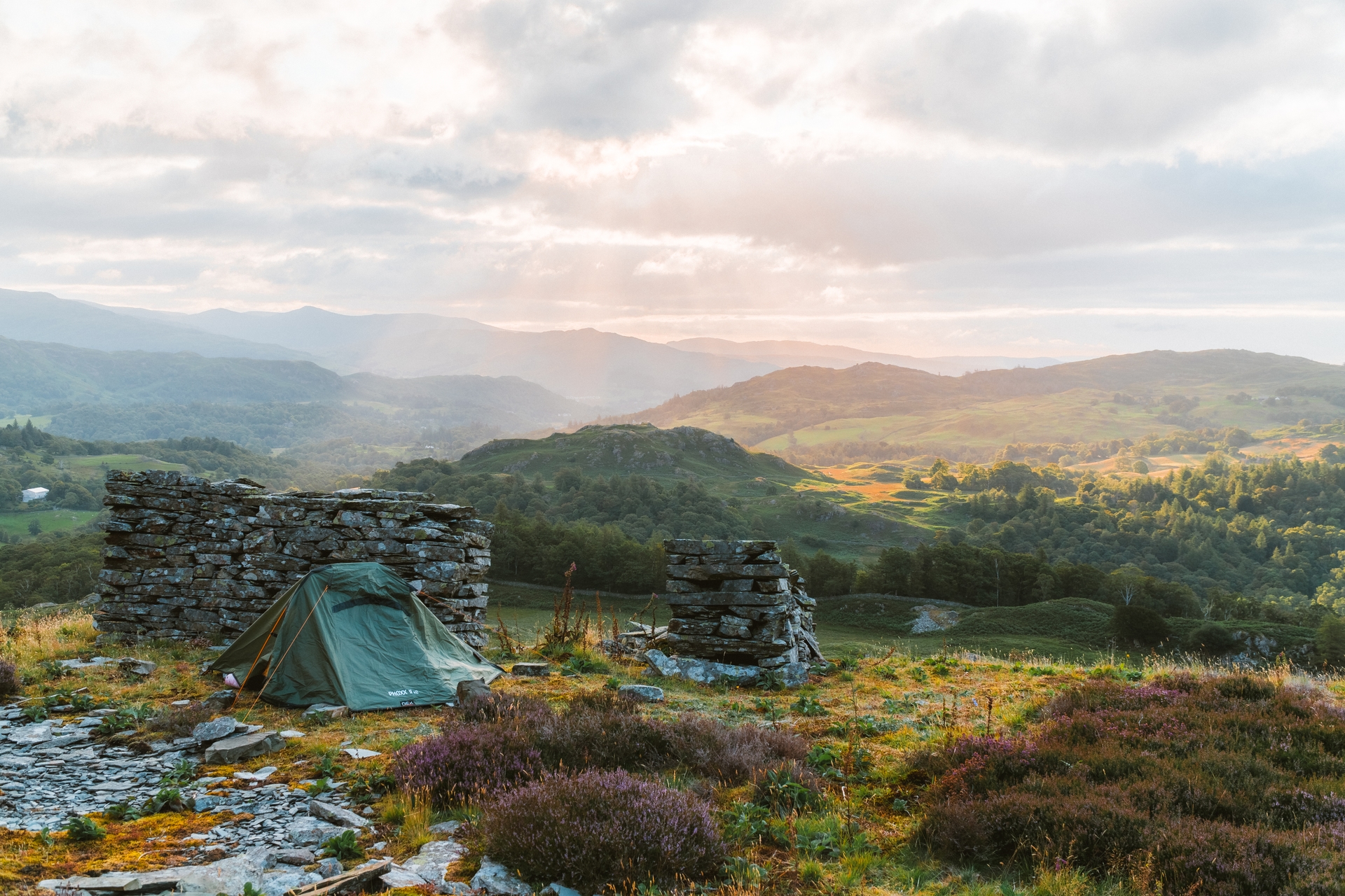 Green tent pitched amidst stone ruins with views of distant mountains and a cloudy sky.