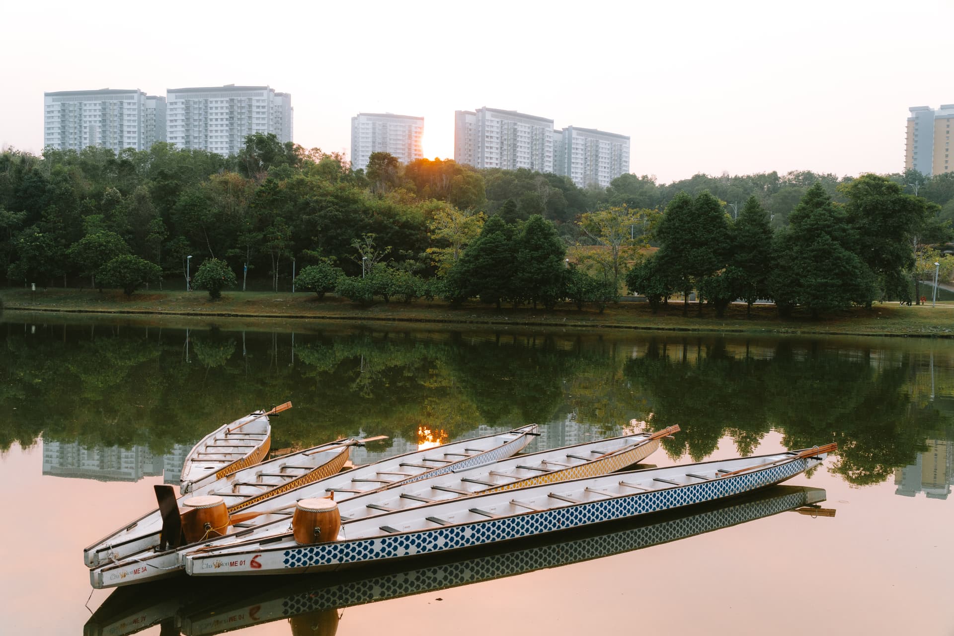 Dragon boats rest on a calm lake, reflecting the morning light and surrounding cityscape.