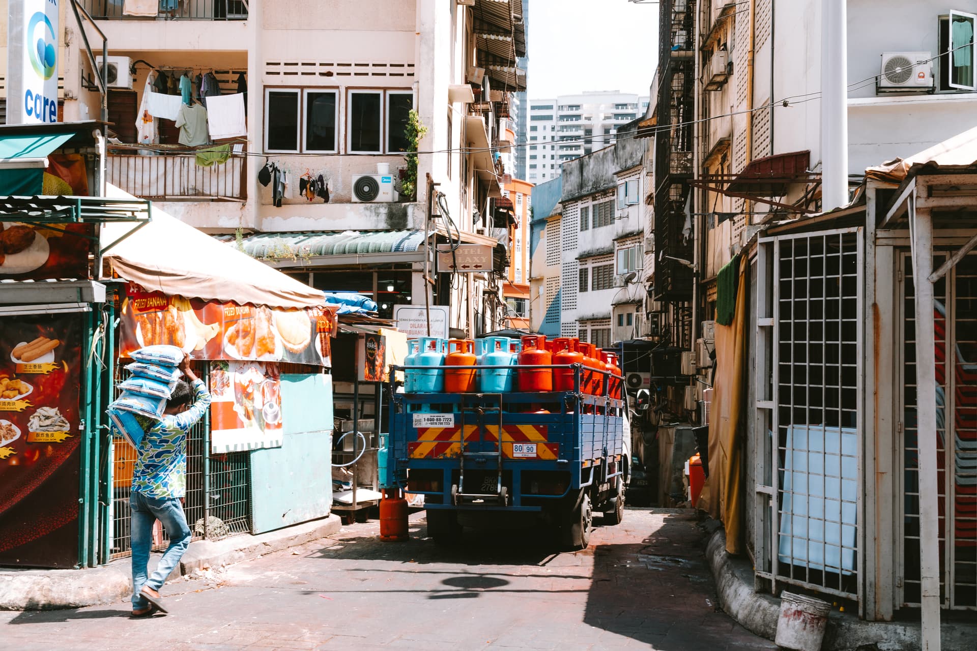 A man carries goods through a bustling city alley, with a truck full of tanks in the background.